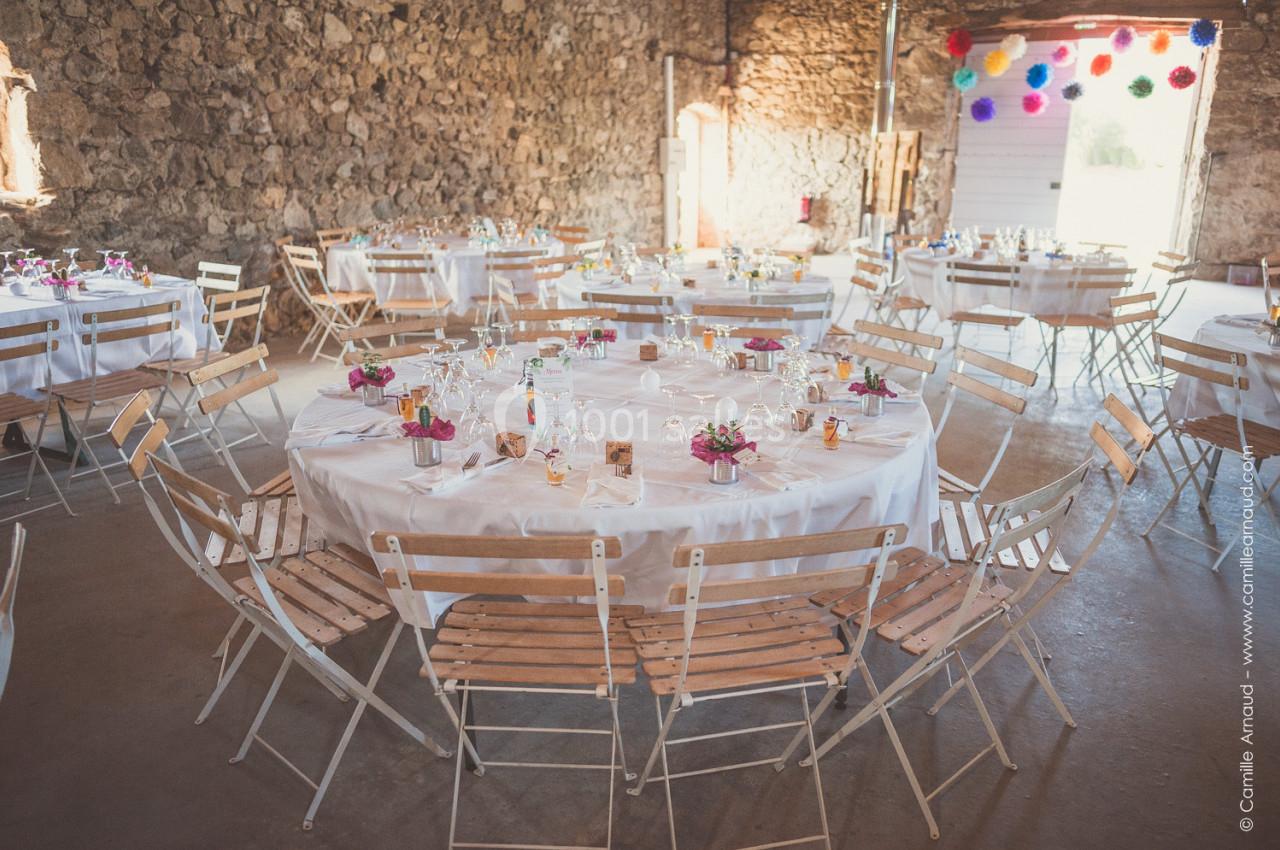Salle de réception rustique avec tables rondes dressées, nappes blanches et décorations colorées suspendues.