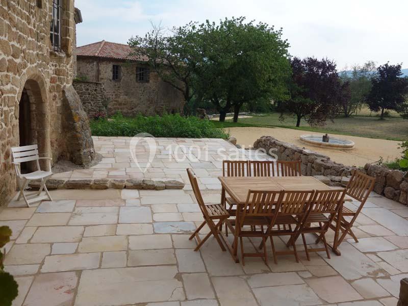 Terrasse en pierre avec table et chaises en bois, entourée de verdure et d'un bâtiment en pierre.