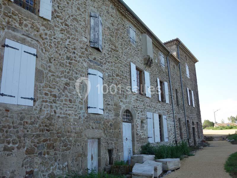 Façade en pierre d'un bâtiment ancien avec volets blancs, chemin en gravier et végétation en bordure.