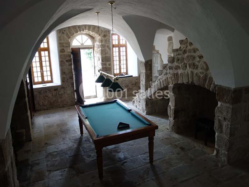 Salle voûtée en pierre avec une table de billard éclairée par des lampes suspendues et des fenêtres en arrière-plan.