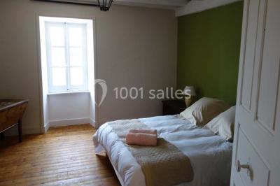 Chambre avec un lit double, draps blancs, couverture rouge, deux oreillers bleus et un mur rouge, éclairée par une fenêtre.