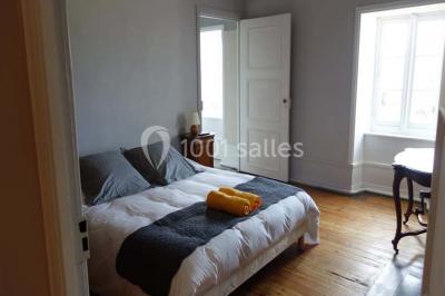 Chambre avec un lit double, draps blancs, couverture rouge, deux oreillers bleus et un mur rouge, éclairée par une fenêtre.