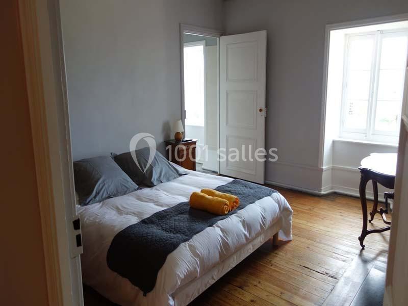 Chambre lumineuse avec un lit double, draps blancs, couverture grise, serviettes jaunes et parquet en bois clair.