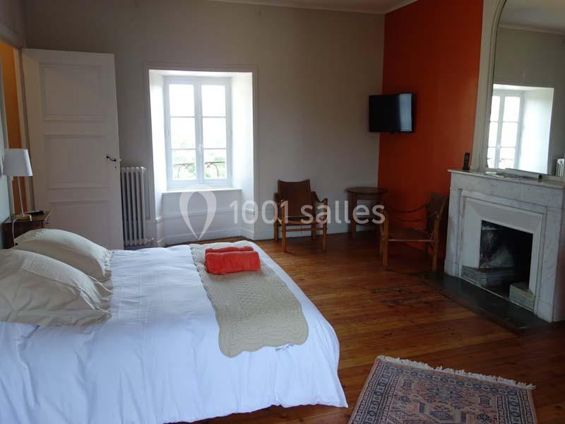 Chambre lumineuse avec lit double, parquet en bois, cheminée, mur orange, télévision murale et grandes fenêtres.