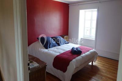 Chambre avec un lit double, draps blancs, couverture rouge, deux oreillers bleus et un mur rouge, éclairée par une fenêtre.