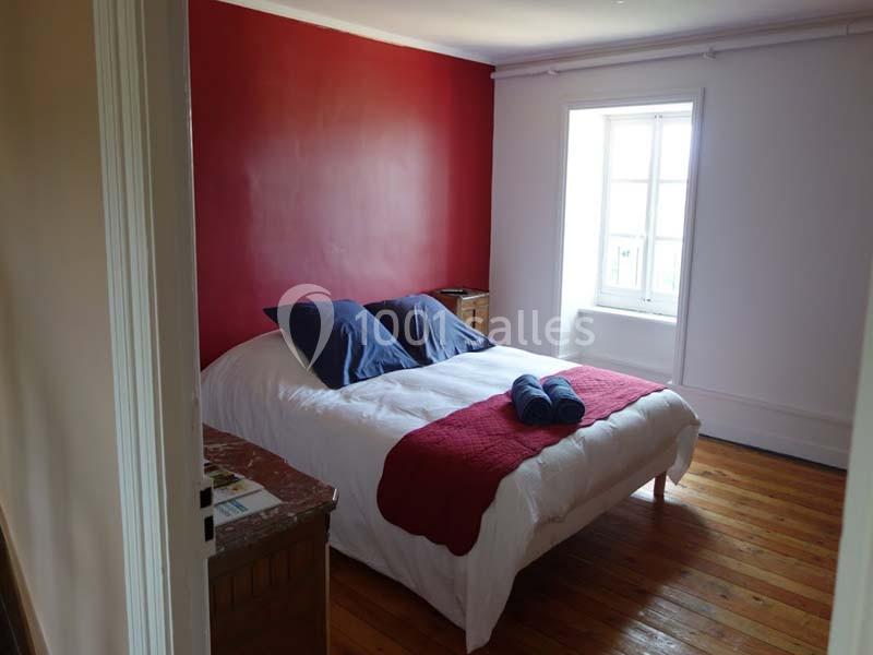 Chambre avec un lit double, draps blancs, couverture rouge, deux oreillers bleus et un mur rouge, éclairée par une fenêtre.