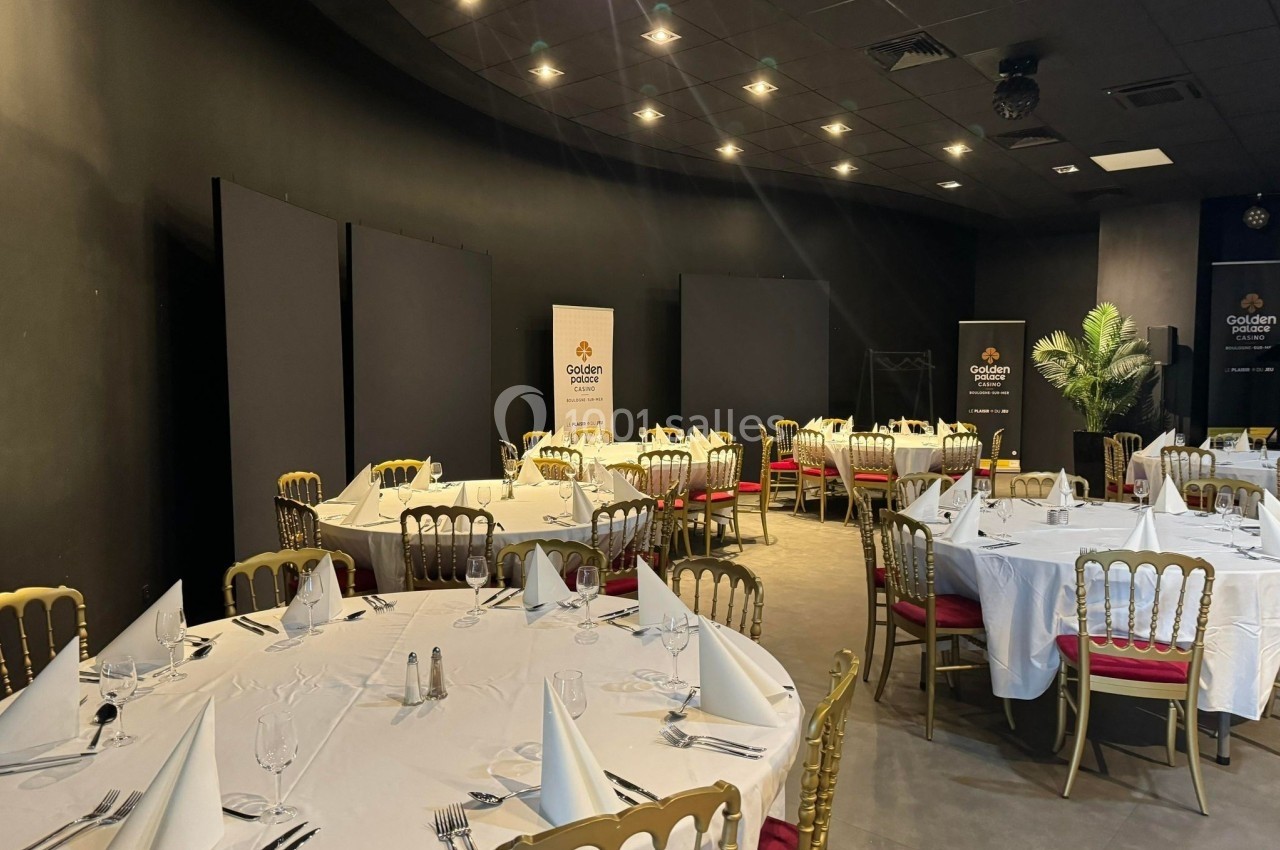 Salle de réception avec tables rondes dressées, nappes blanches, chaises dorées et éclairage tamisé.