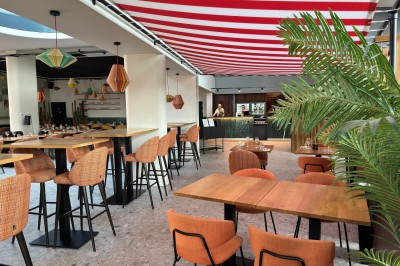 Salle de restaurant lumineuse avec tables en bois, chaises orange et plafond décoré d'un auvent rayé rouge et blanc.