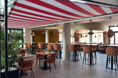 Salle de restaurant lumineuse avec tables en bois, chaises orange et plafond décoré d'un auvent rayé rouge et blanc.