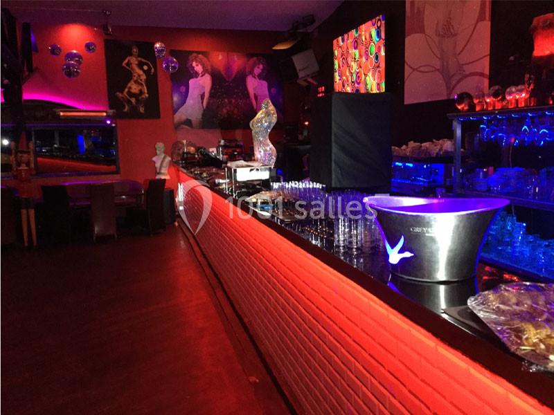 Bar illuminé avec des néons rouges, comptoir long avec verres alignés, décorations artistiques et ambiance tamisée.