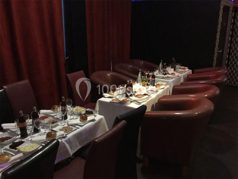 Tables dressées avec nappes blanches, couverts, verres, boissons et plats, entourées de fauteuils rouges dans une salle…