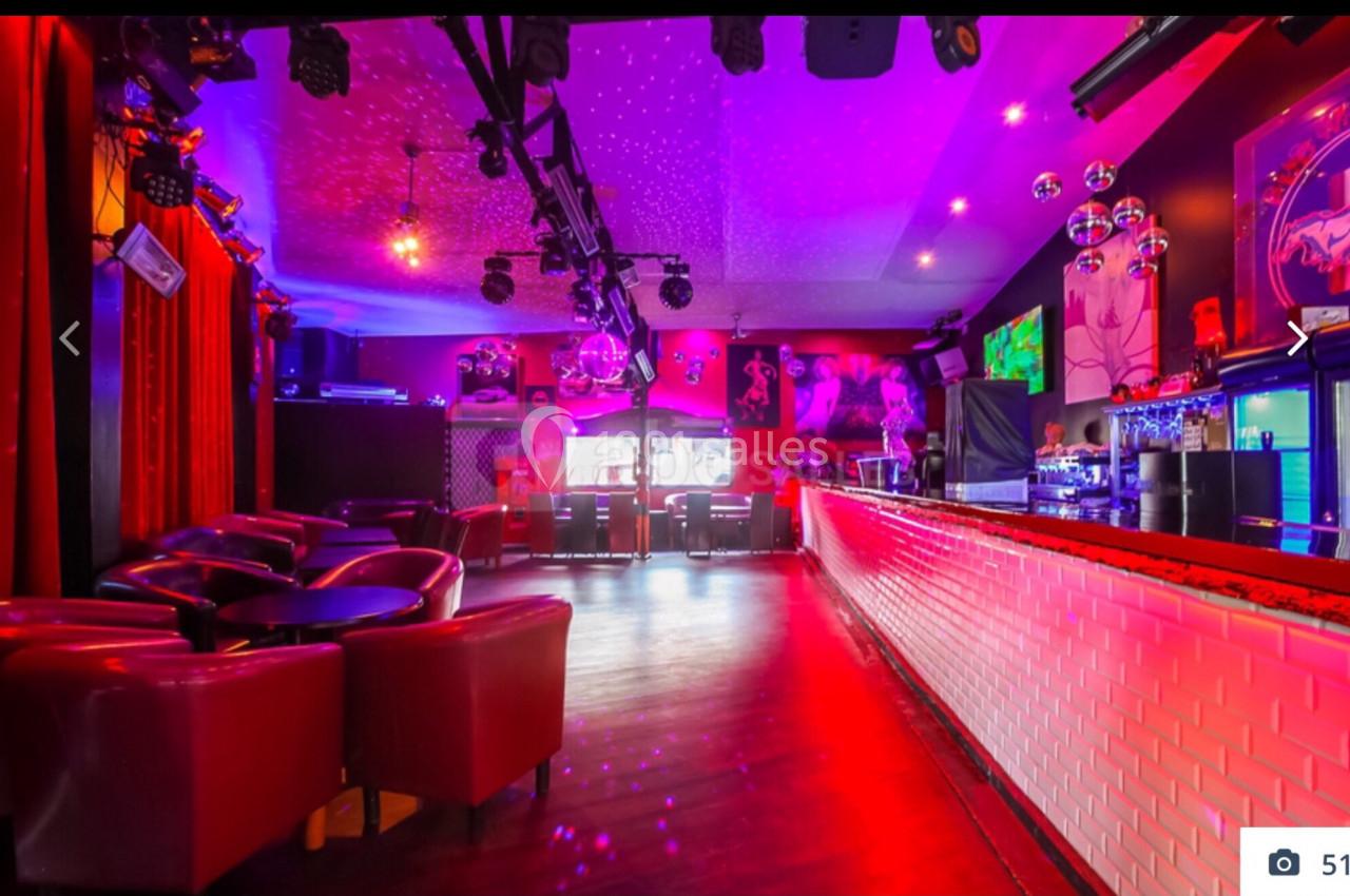 Salle de bar avec éclairage coloré, fauteuils rouges, long comptoir et décorations murales lumineuses.