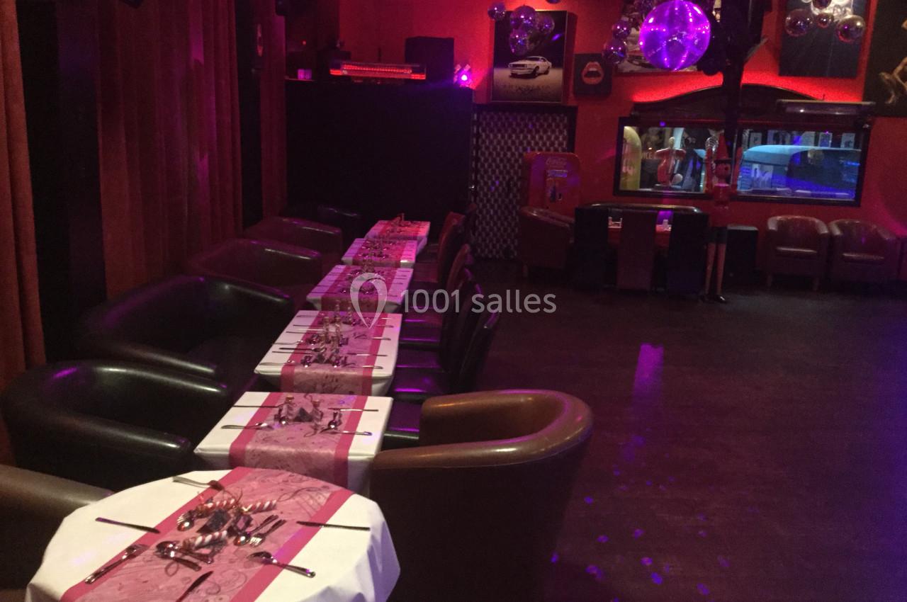 Salle décorée avec des tables alignées, nappes roses et couverts, éclairage tamisé et ambiance festive.