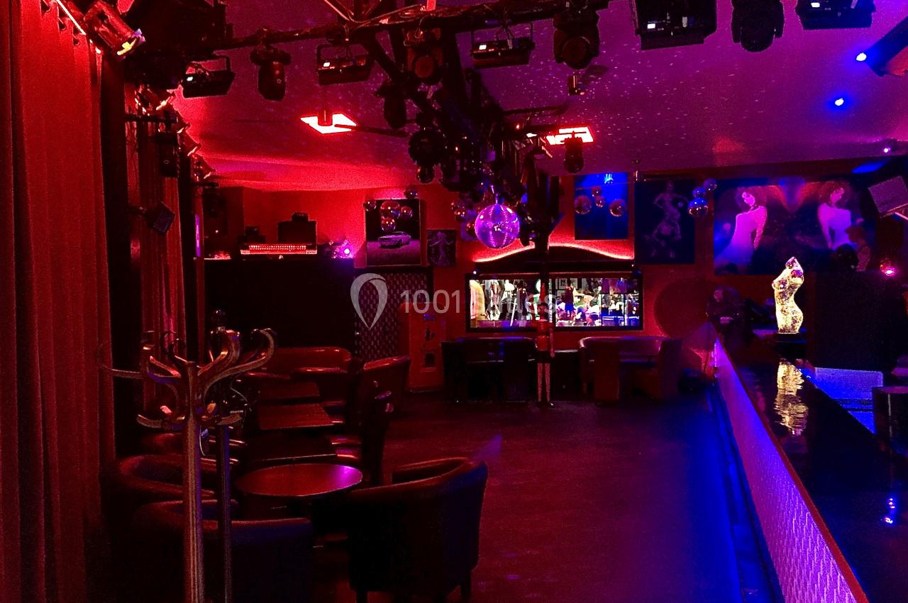 Salle de bar ou discothèque avec éclairage rouge et bleu, tables, fauteuils et bar en arrière-plan.