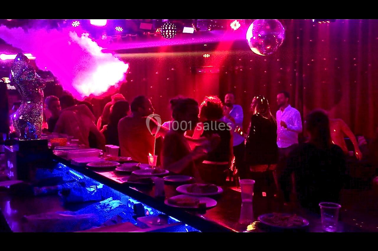 Personnes dansant et discutant dans une ambiance festive avec éclairages colorés, bar au premier plan.