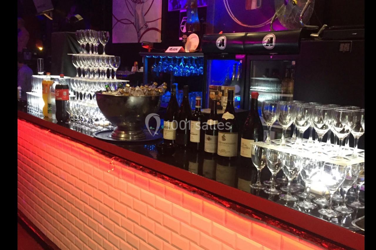 Bar illuminé avec des verres à vin alignés, des bouteilles et une fontaine à champagne sur le comptoir.