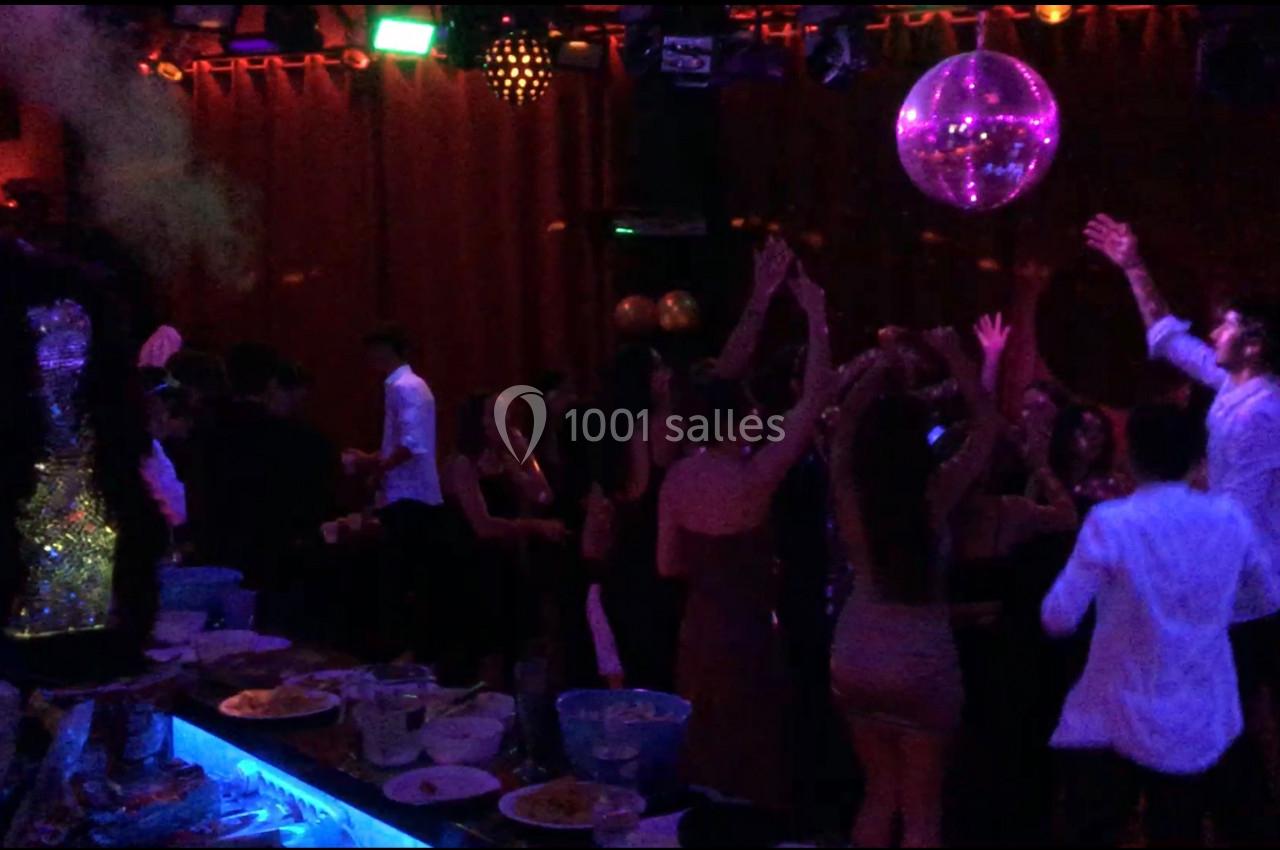 Des personnes dansent sous une boule disco dans une ambiance de club avec des lumières colorées et un bar en premier plan.