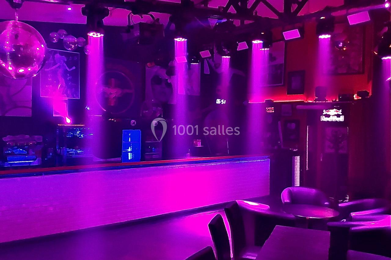 Salle de bar ou discothèque éclairée par des lumières violettes, avec tables, chaises et un comptoir en arrière-plan.