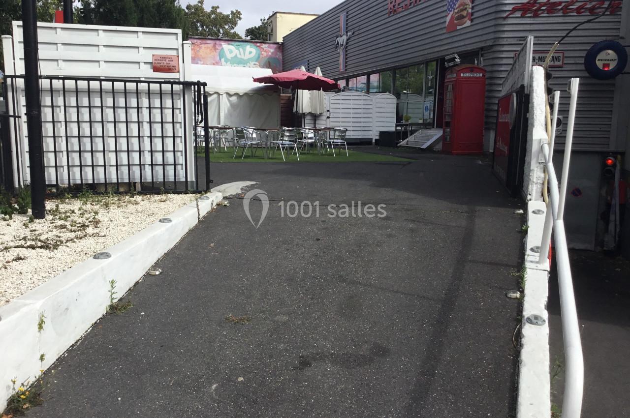 Allée asphaltée menant à une terrasse extérieure avec tables, chaises et parasols, près d'un bâtiment commercial.