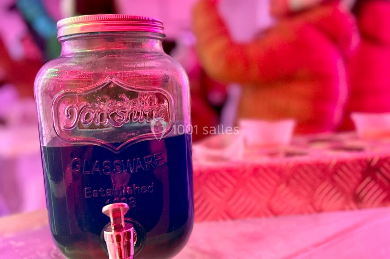 Bocal en verre rempli de liquide bleu avec un robinet, posé sur une surface glacée dans un environnement lumineux rose.