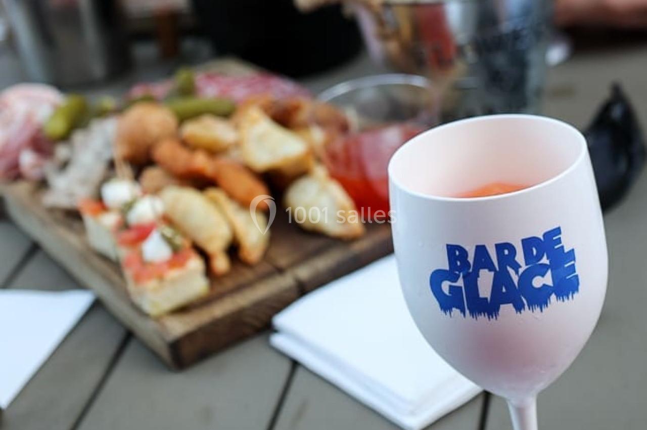Verre de boisson marqué ’Bar de Glace’ au premier plan, plateau de bouchées variées flou en arrière-plan.