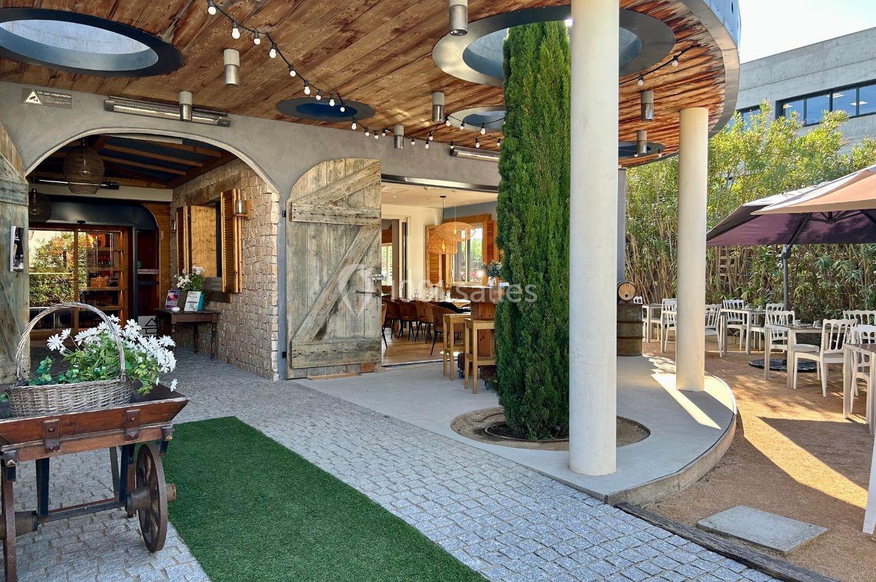 Entrée d'un restaurant avec terrasse extérieure, mobilier en bois, plantes et éclairage suspendu sous un toit en bois.