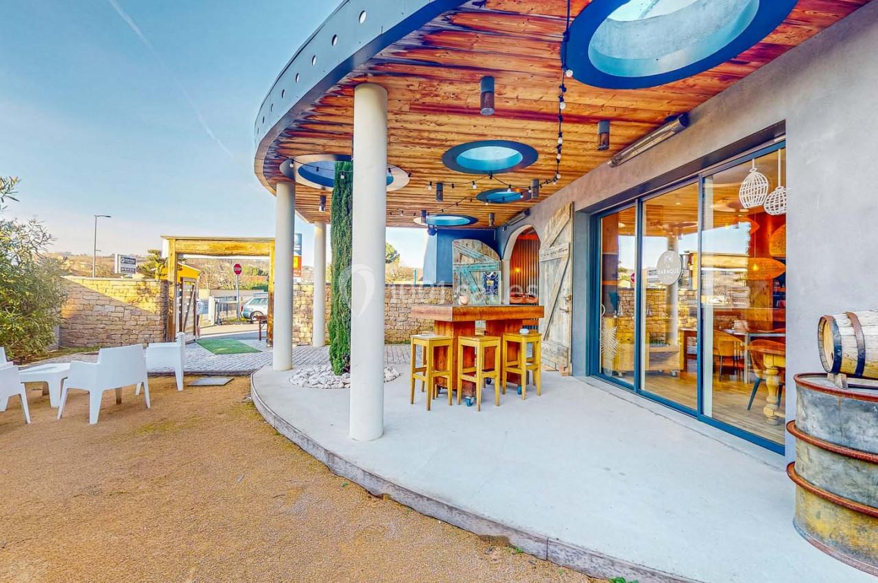 Terrasse extérieure avec tables en bois, chaises blanches, tonneaux décoratifs et toit circulaire ajouré.
