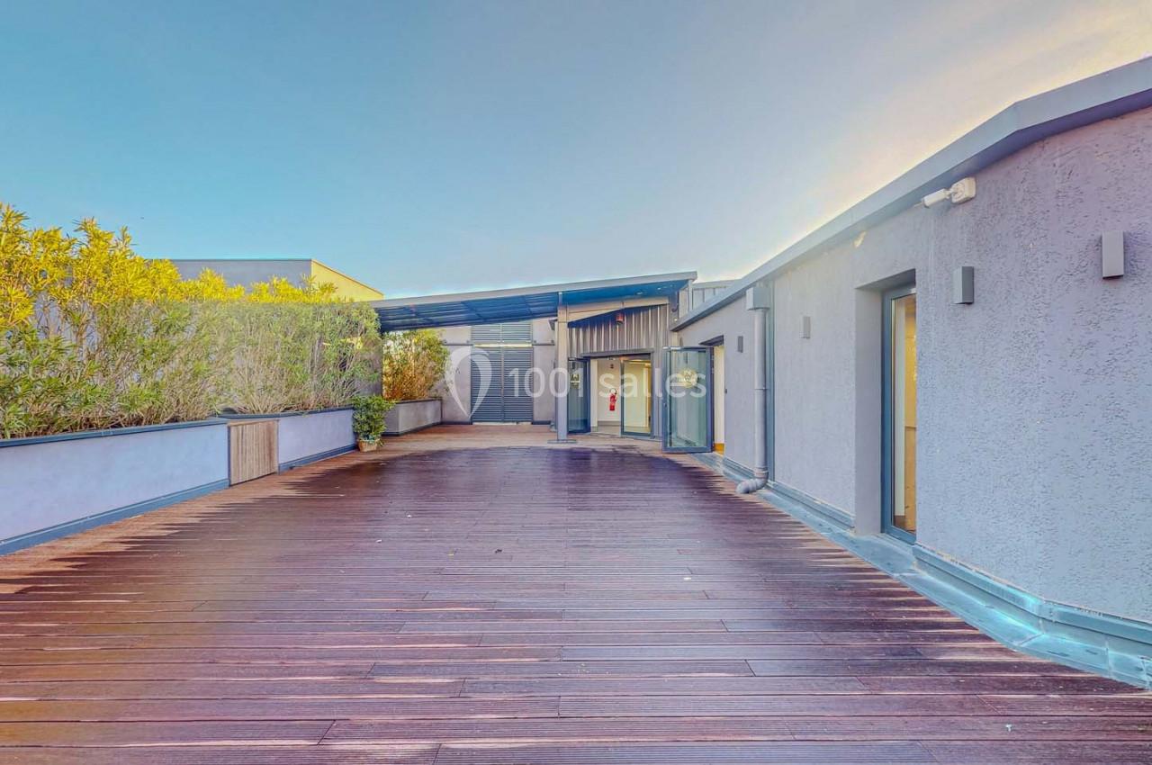 Terrasse en bois avec végétation sur le côté, entourée de bâtiments modernes sous un ciel dégagé.