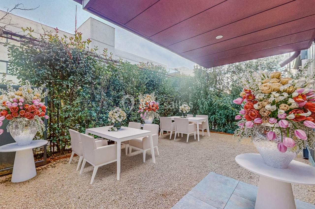 Terrasse extérieure avec tables blanches, chaises assorties et arrangements floraux colorés, entourée de végétation.