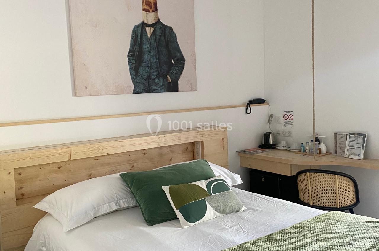 Chambre lumineuse avec lit double, tête de lit en bois, plaid vert, tableau mural et coin bureau avec chaise en rotin.