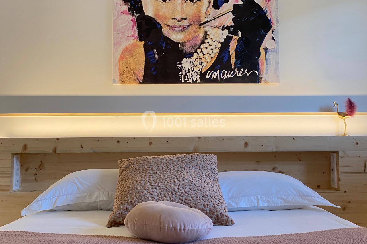 Lit double avec couverture rose, coussins assortis et tableau coloré représentant un visage féminin au mur.