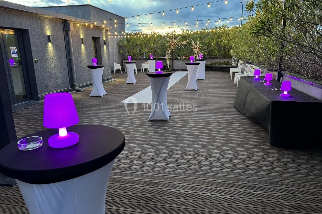Terrasse en bois aménagée avec des tables hautes, lampes violettes allumées et guirlandes lumineuses suspendues.