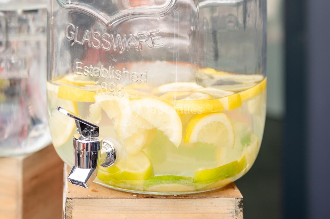 Distributeur en verre rempli d'eau infusée avec des tranches de citron et de citron vert, posé sur un support en bois.