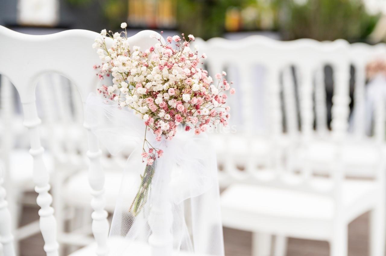 Bouquet de gypsophiles et fleurs roses attaché à une chaise blanche lors d'une cérémonie en extérieur.