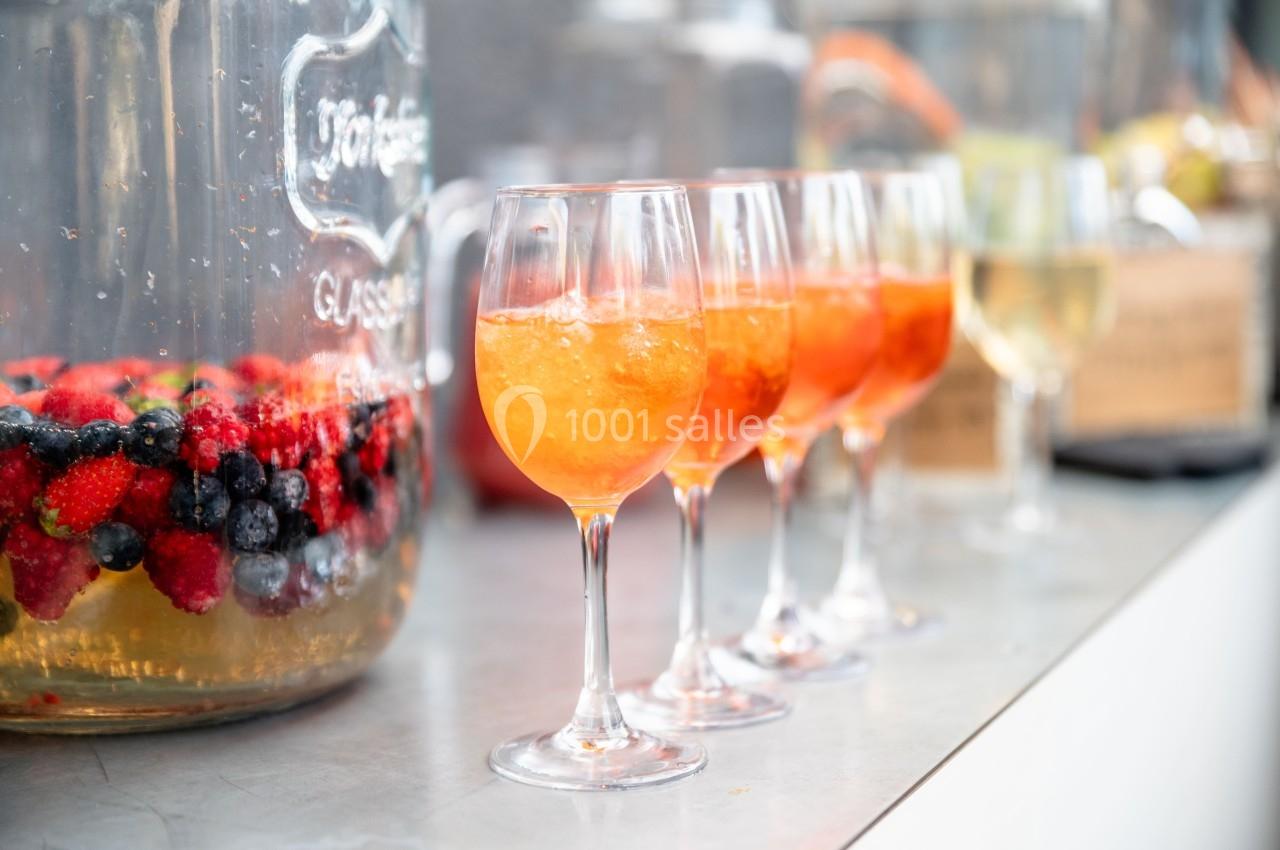 Quatre verres de cocktail orange alignés sur un comptoir, avec une jarre de fruits rouges en arrière-plan.