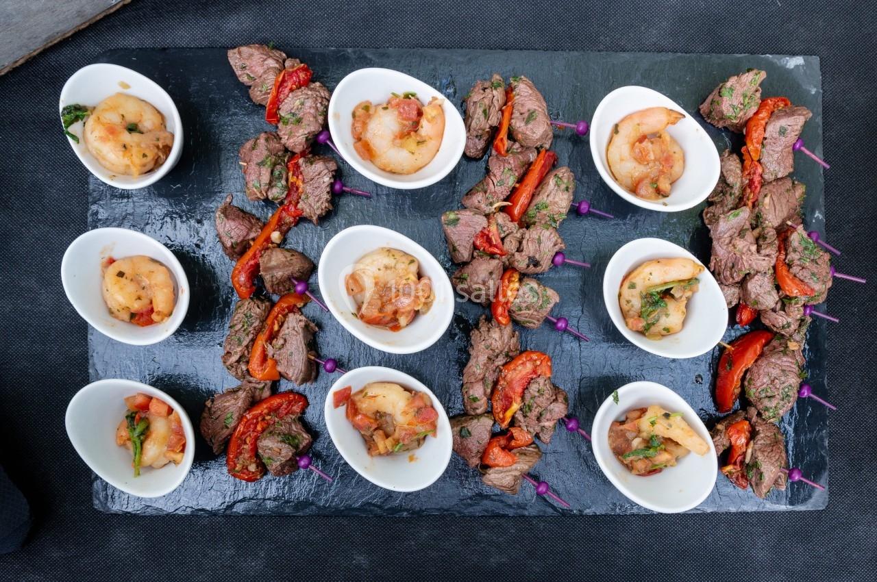 Assortiment de brochettes de viande et crevettes marinées, accompagné de bouchées servies dans des coupelles blanches.