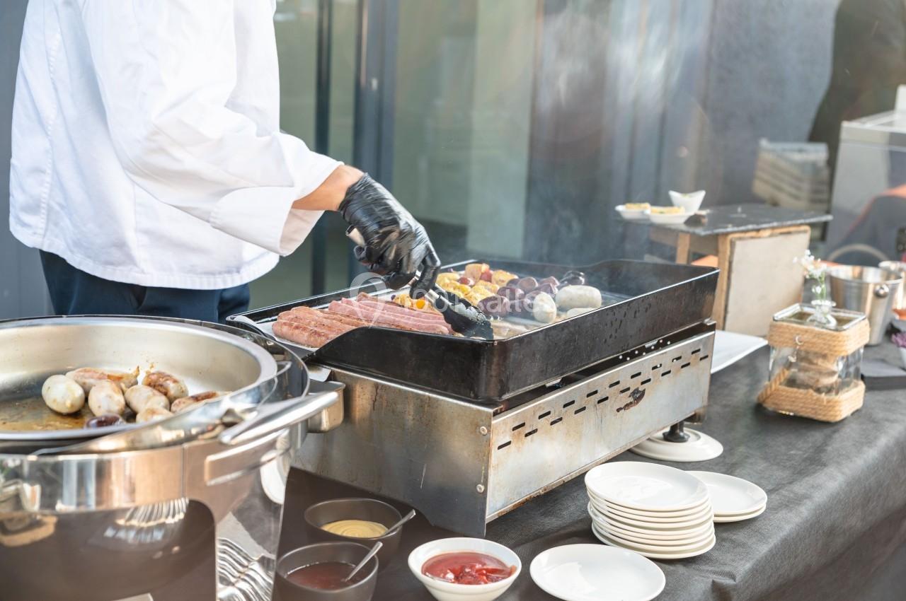 Un cuisinier en tenue blanche grille des saucisses et légumes sur une plancha lors d'un service en extérieur.