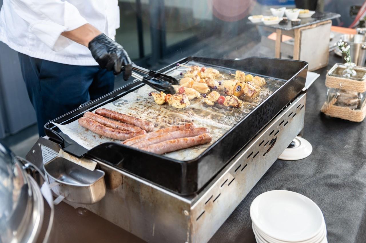Un cuisinier prépare des saucisses et des légumes grillés sur une grande plancha en extérieur.