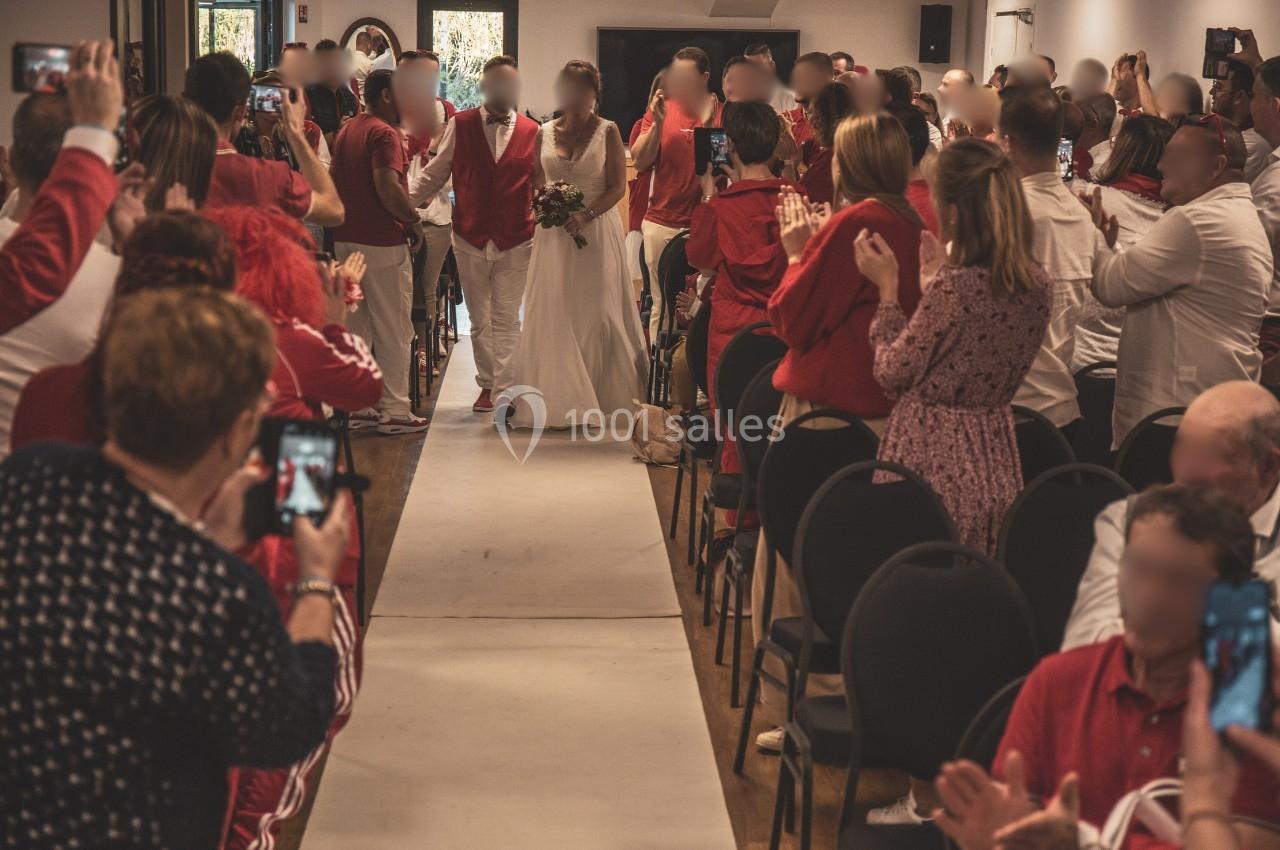 Un couple en tenue de mariage marche dans l'allée centrale d'une salle, entouré d'invités applaudissant.