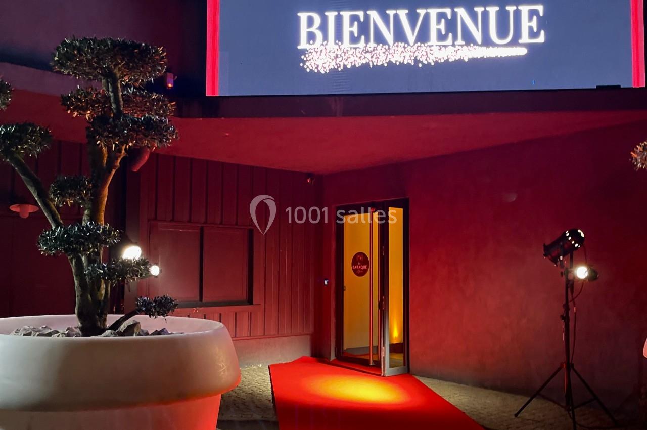 Entrée éclairée avec tapis rouge, panneau ’Bienvenue’ lumineux et plante décorative dans un pot blanc.
