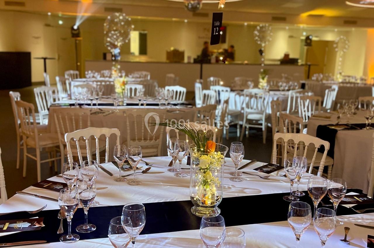 Salle de réception décorée avec des tables rondes dressées, centre de table floral et ballons suspendus.
