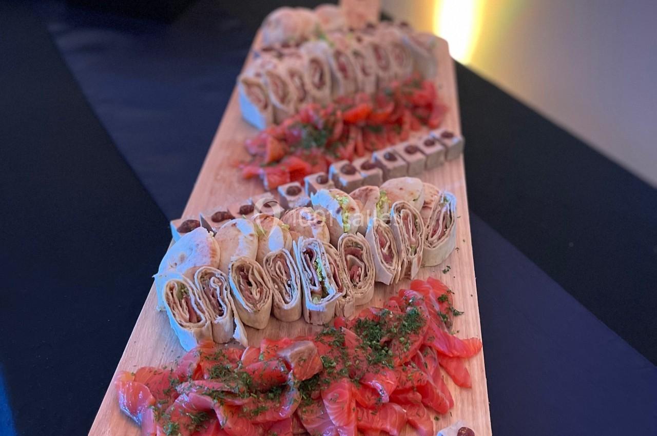 Plateau en bois avec des roulés apéritifs, du saumon fumé et des bouchées sucrées, présenté sur une table noire.
