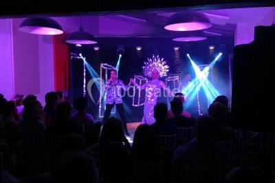 Un artiste chante sur scène avec une personne en costume étincelant sous des lumières violettes, devant un public assis.