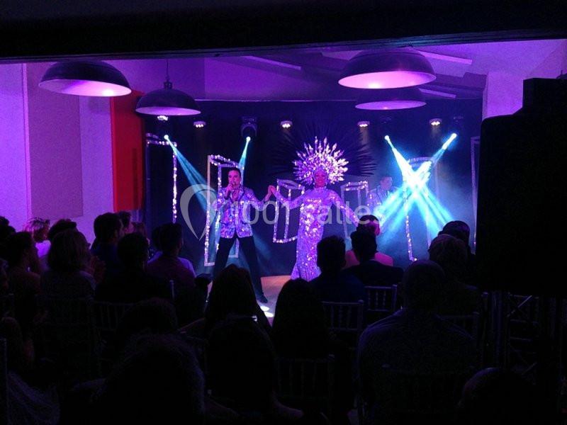 Un artiste chante sur scène avec une personne en costume étincelant sous des lumières violettes, devant un public assis.