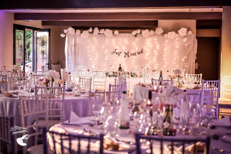 Salle de réception décorée pour un mariage avec des tables dressées et un fond lumineux orné de fleurs blanches.