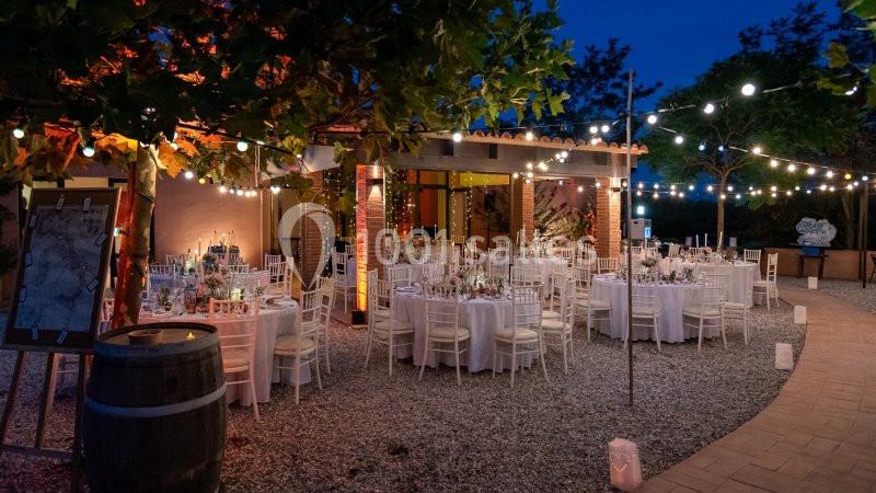 Terrasse éclairée en soirée avec des tables rondes dressées pour un événement, entourées de guirlandes lumineuses.
