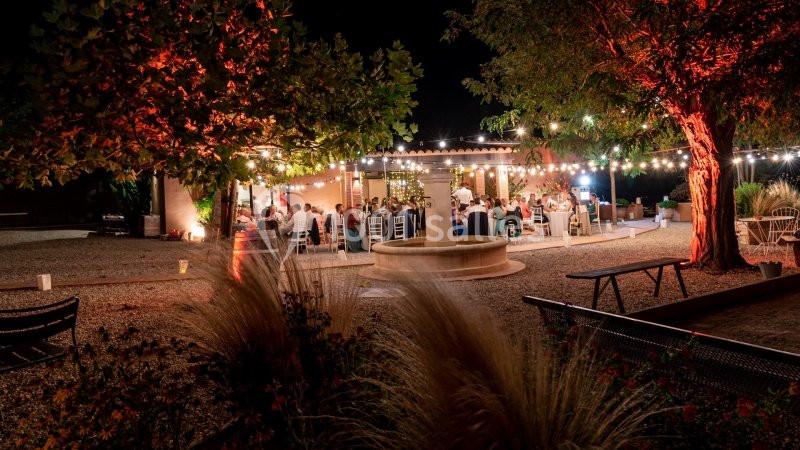 Terrasse extérieure éclairée par des guirlandes lumineuses, avec des invités dînant sous les arbres en soirée.