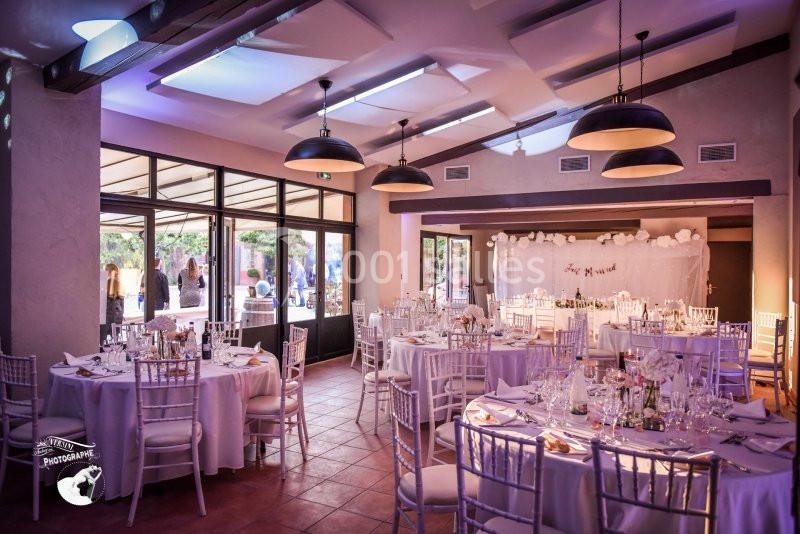 Salle de réception décorée pour un mariage, avec tables rondes dressées, chaises blanches et éclairage chaleureux.