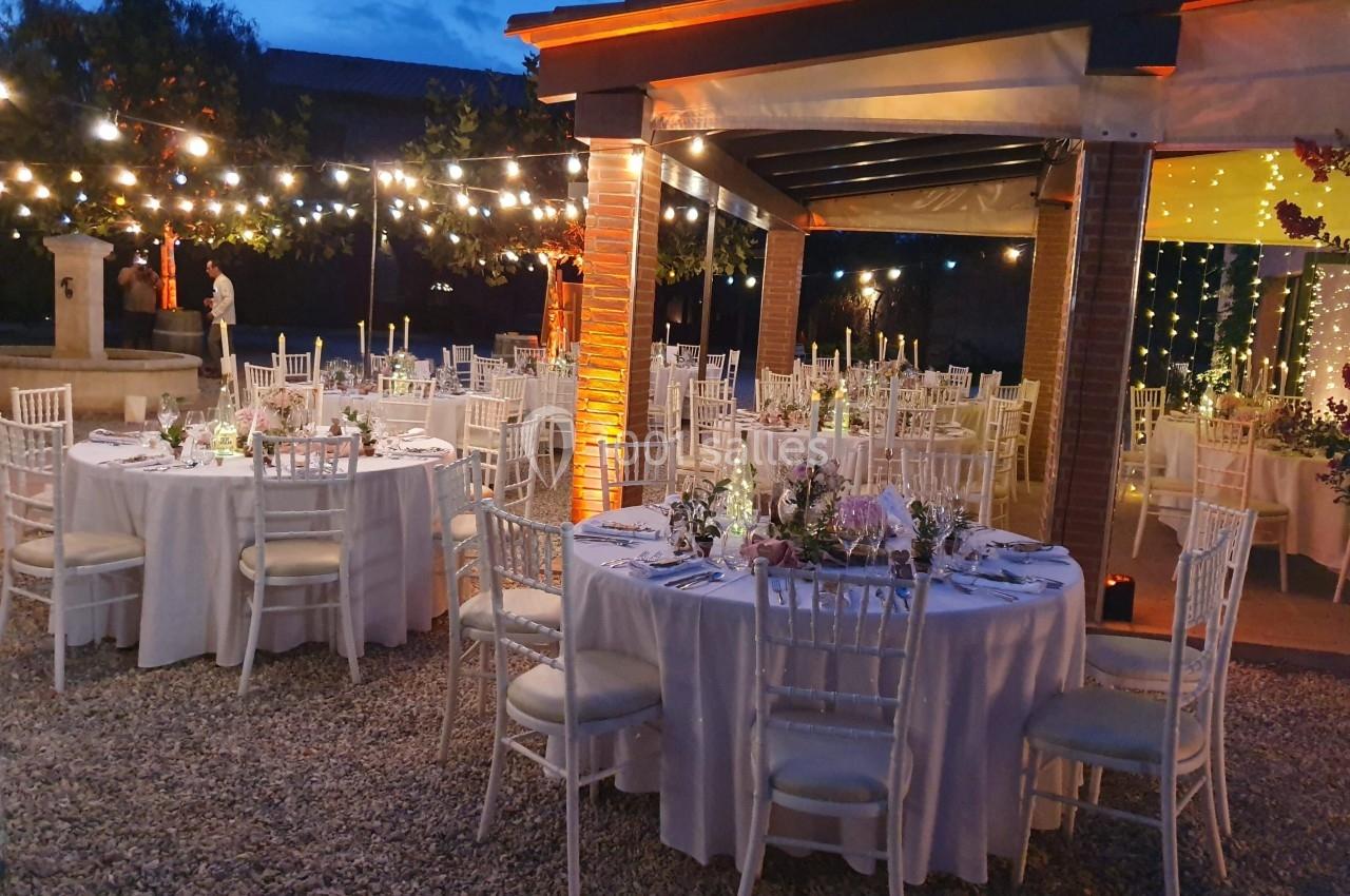 Tables rondes décorées pour un événement en soirée, disposées sous une pergola éclairée par des guirlandes lumineuses.