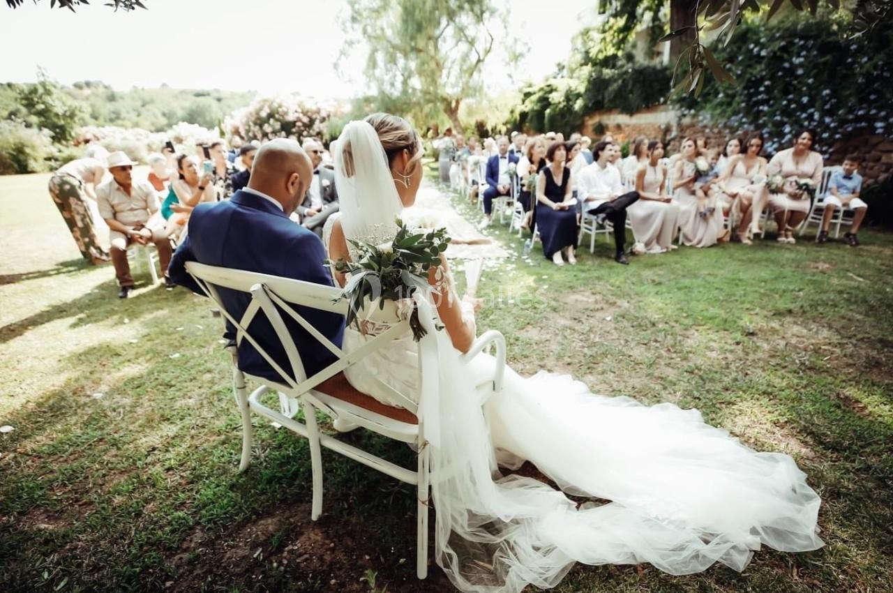 Un couple de mariés assis de dos lors d'une cérémonie en plein air, entouré d'invités sur des chaises.